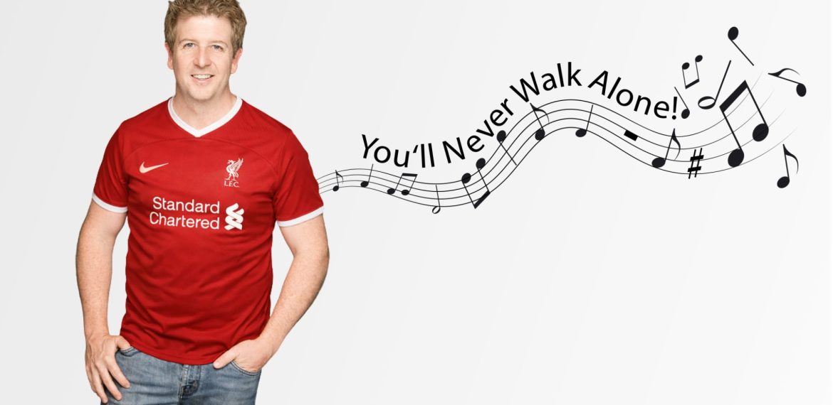 A smiling man in a red Liverpool football jersey stands beside musical notes and the text “You’ll Never Walk Alone!” flowing on a staff across a light background, echoing the uplifting vibe of Hot 1027 Breakfast.