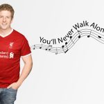 A smiling man in a red Liverpool football jersey stands beside musical notes and the text “You’ll Never Walk Alone!” flowing on a staff across a light background, echoing the uplifting vibe of Hot 1027 Breakfast.