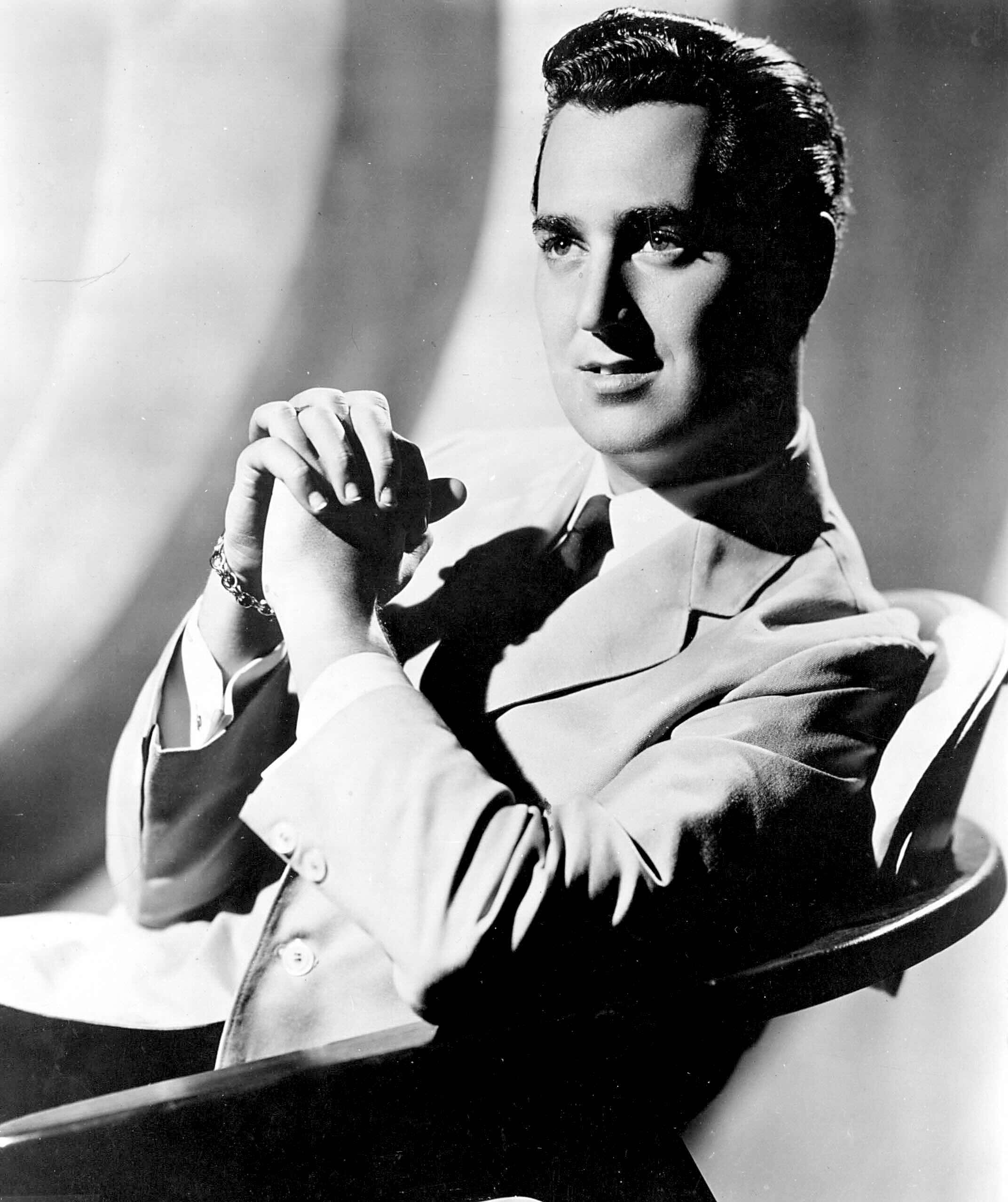 Black and white portrait of a man in a light-colored suit, sitting in a chair with his hands clasped, gazing slightly to the side with a calm expression. Soft lighting creates gentle shadows, as smooth as blancmange, in the background.