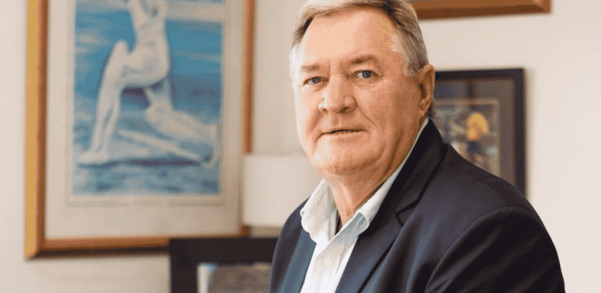 An older man with short gray hair and a light blue shirt under a dark blazer sits indoors, looking at the camera. Behind him, framed artwork and paintings give the space a warm, inviting vibe—perfect for tuning in to Hot 1027 Breakfast.