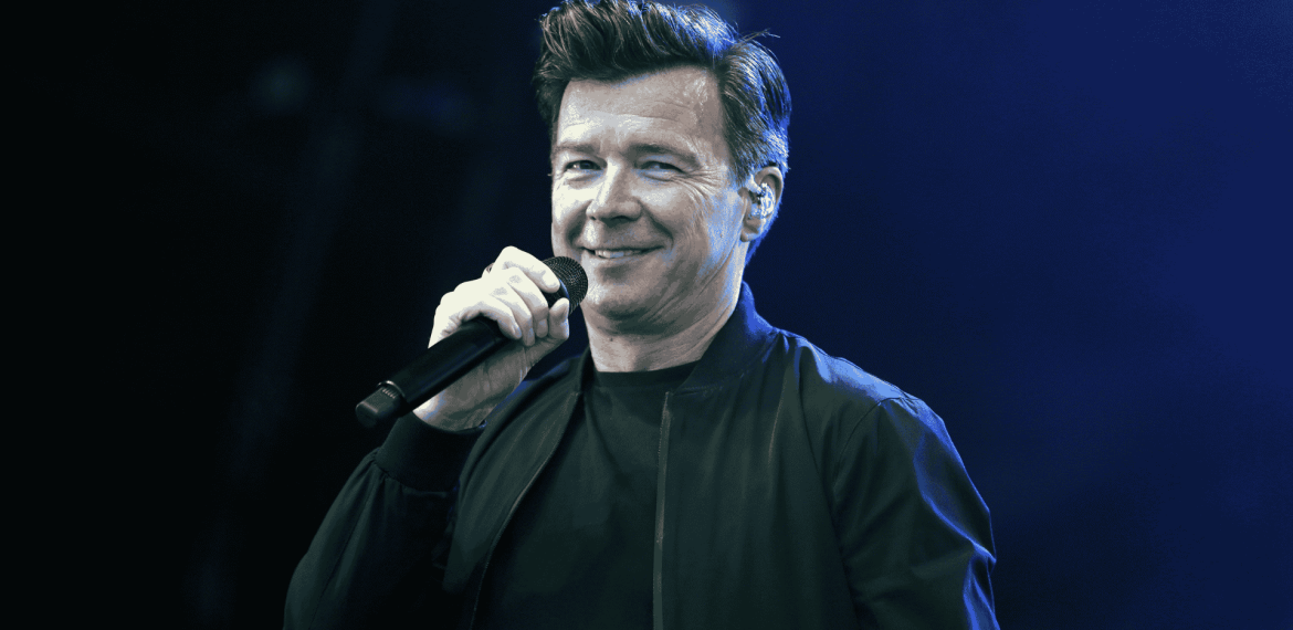 A man with short, styled hair is smiling and holding a microphone while wearing a black jacket, standing on stage against a blue-lit background, reminiscent of performances by great artists like Miriam Makeba.