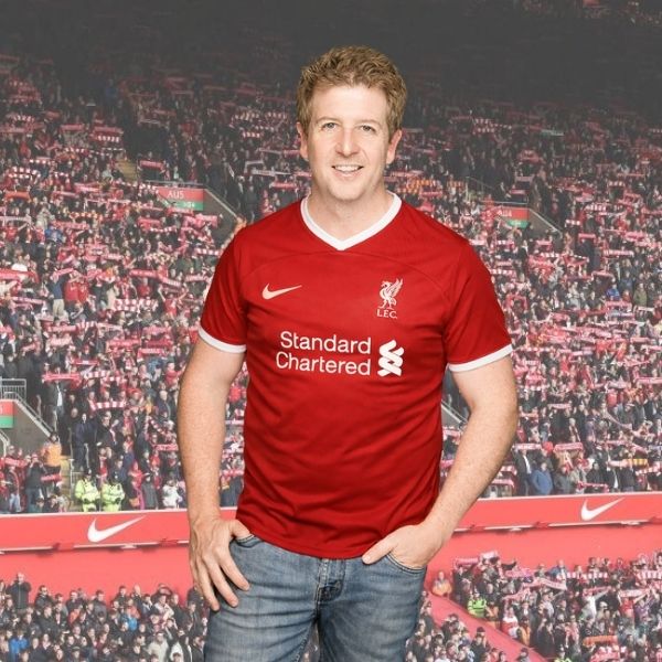A smiling man in a red Liverpool FC soccer jersey stands in front of a stadium filled with fans holding red and white scarves, ready to cheer as if they’re tuning in to the excitement of the Hot 1027 Breakfast show.