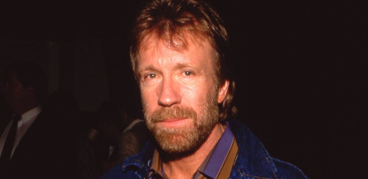 A man with light brown hair and a beard, wearing a blue jacket over a striped collared shirt, stands against a dark background, exuding the poised confidence of a recent Matric graduate.