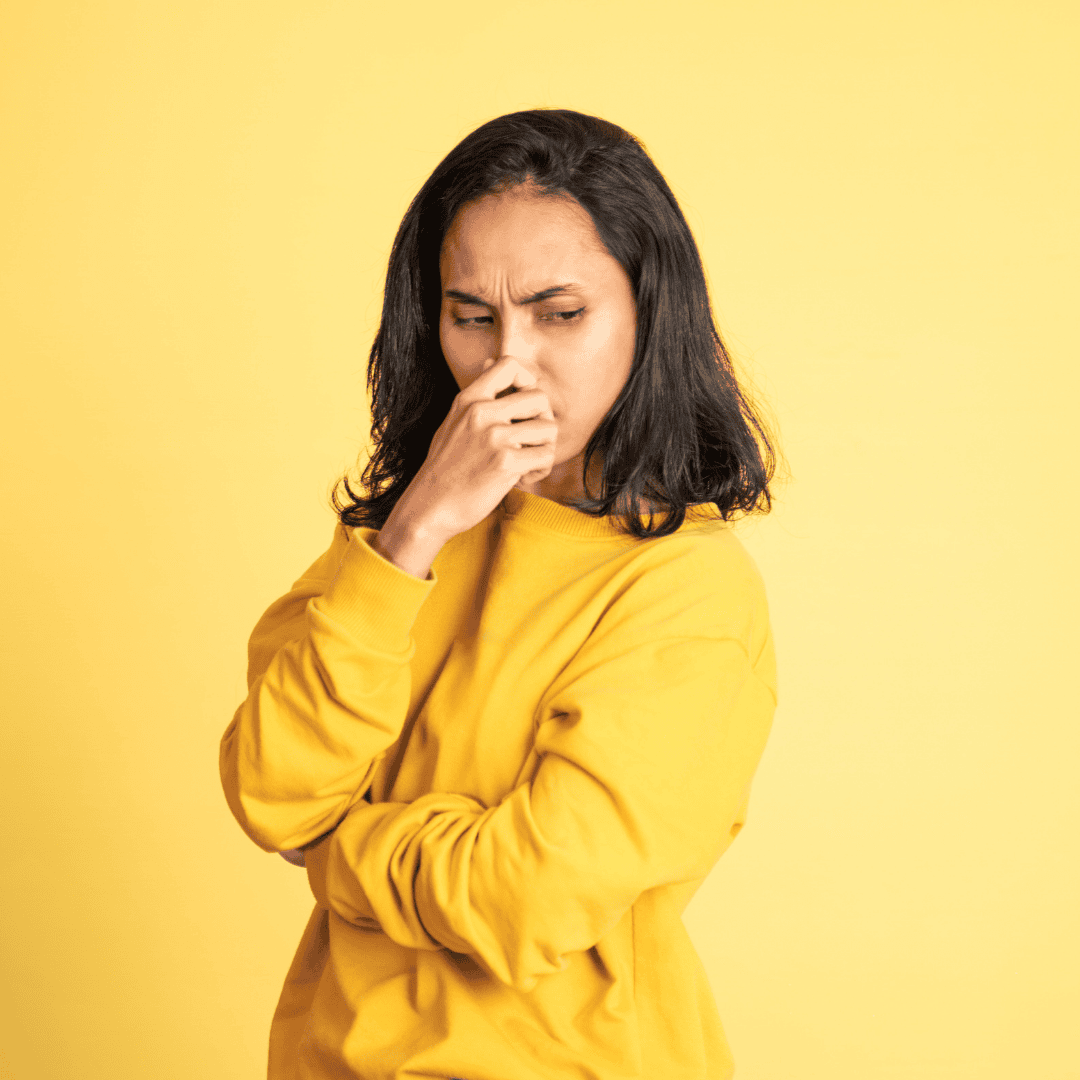 A person in a yellow sweater stands against a yellow background, holding their nose and looking displeased, as if the hot 1027 breakfast nearby has an unpleasant smell.