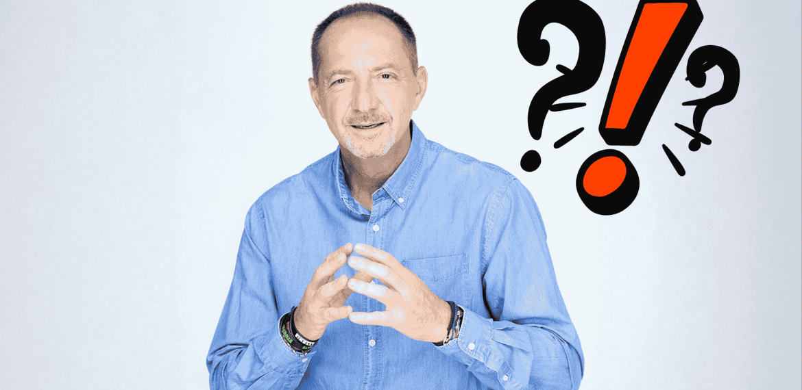 A man in a blue shirt gestures with his hands and smiles at the camera. Behind him are bold question marks and an exclamation mark, suggesting travel confusion or surprise. The background is light and plain.