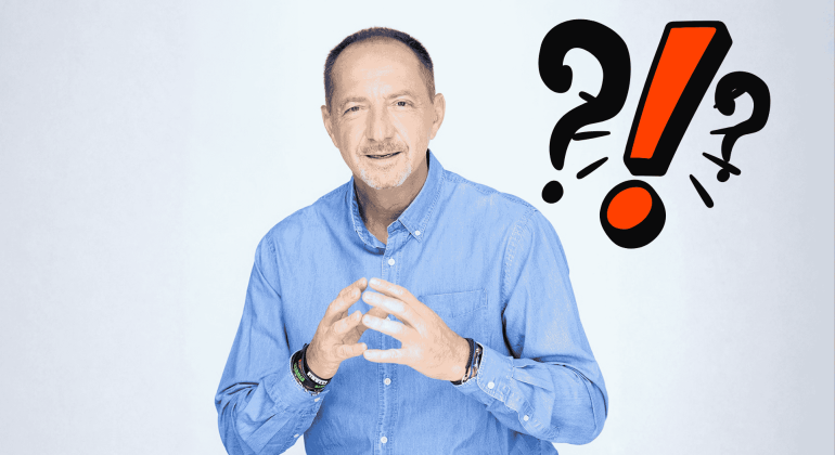 A man in a blue shirt gestures with his hands and smiles at the camera. Behind him are bold question marks and an exclamation mark, suggesting travel confusion or surprise. The background is light and plain.