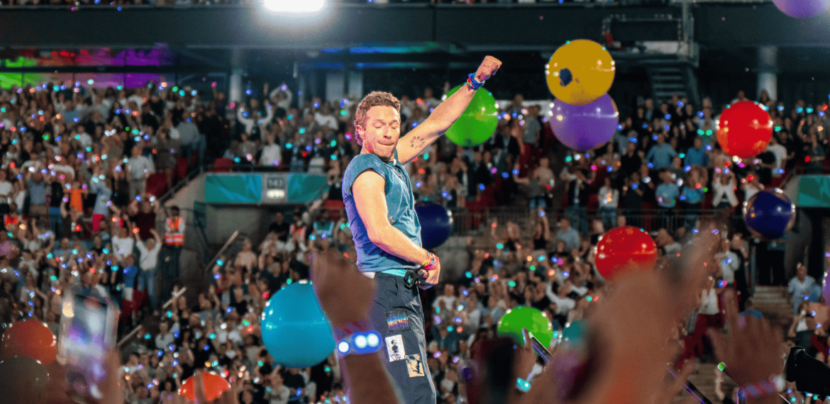 A musician performs energetically on stage in front of a large, cheering crowd at the Hot 1027 Breakfast, with colorful balloons floating and audience members raising their hands and phones.