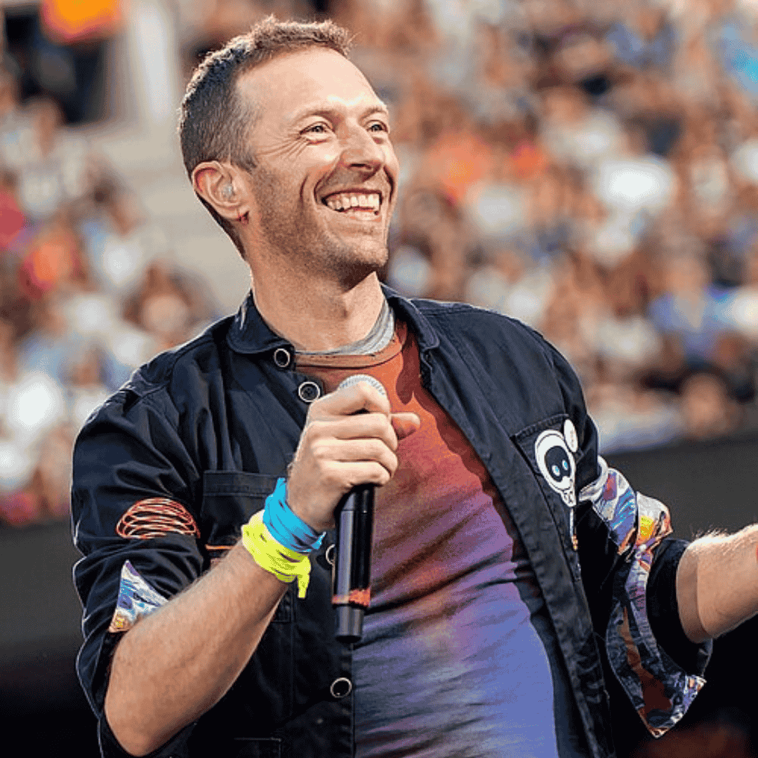 A smiling man wearing a dark jacket with colorful patches, holding a microphone, stands on stage in front of a blurred crowd at the Hot 1027 Breakfast outdoor event.
