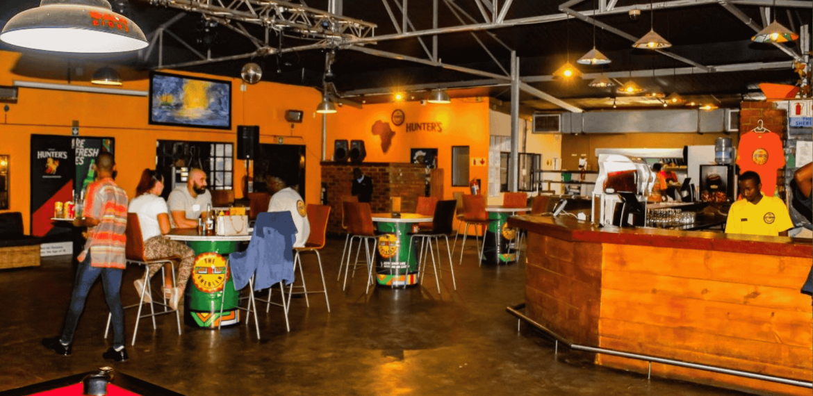 A brightly lit bar with orange walls, scattered tables and chairs, people socializing over hot 1027 breakfast, two TV screens, a pool table in the foreground, and a wooden counter on the right.
