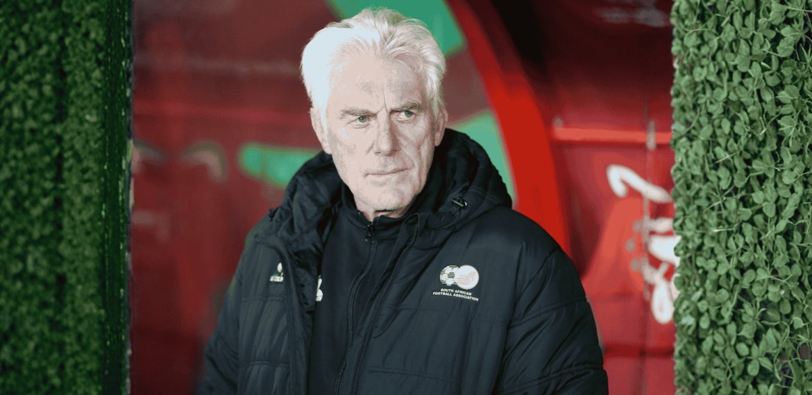 An older man with white hair in a dark jacket stands before red and green colors, leafy green bush walls on either side. With a serious expression, he looks slightly to the side, as if contemplating his next travel getaway.