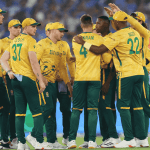 A group of South African cricket players in yellow and green uniforms huddle together on the field during a match, with a crowd visible in the blurred background, capturing the spirit much like a lively Hot 1027 Breakfast gathering.