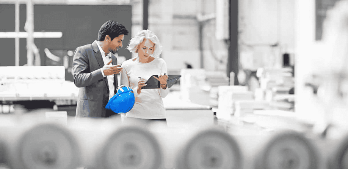 Two professionals, one holding a blue hard hat, stand in a bright industrial setting. As they discuss plans over a tablet, the energy feels as lively as a Hot 1027 Breakfast, with machinery and blurred equipment in the foreground.