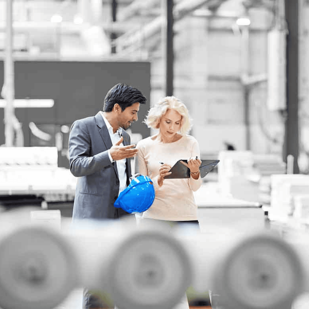 Two professionals, one holding a blue hard hat and the other using a digital tablet, discuss something in an industrial setting, perhaps planning the day's work after listening to Hot 1027 Breakfast with machinery and equipment in the background.