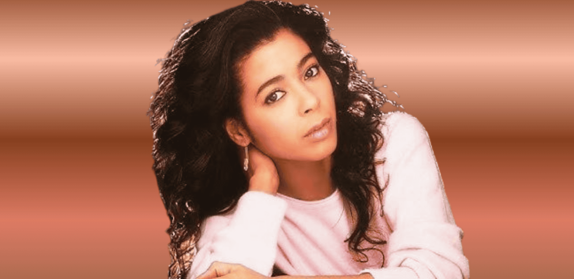 A woman with long, curly dark hair and a light pink sweater rests her head on one hand, looking calmly at the camera against a soft brown gradient background—perfectly capturing the relaxed vibe of a Hot 1027 Breakfast morning.