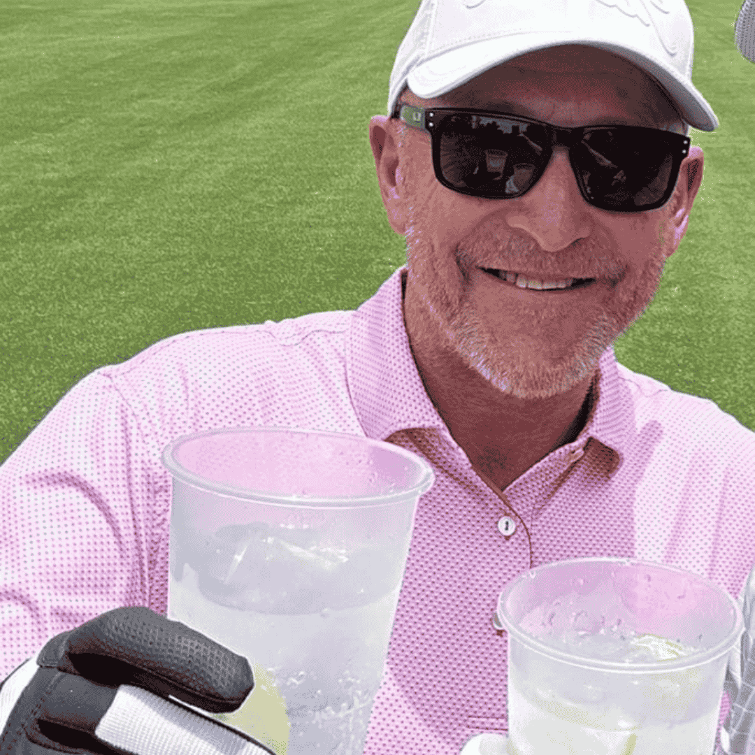 A man in a pink polo shirt, sunglasses, and a white cap smiles while holding a plastic cup with a drink on a green golf course—perfect for a relaxing travel getaway. Another cup and gloved hand are partially visible in the foreground.