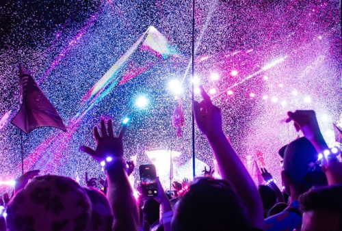 A vibrant crowd at a music festival raises their hands under bright, colorful lights and falling confetti—an unforgettable travel getaway with glowing wristbands and flags waving in the air.