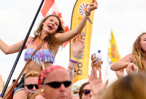 A young woman wearing a fringed top smiles and raises her arms while sitting on someone’s shoulders at a lively outdoor travel getaway. Colorful flags and a crowd of people enjoying the event are visible in the background.