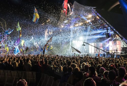 A large crowd at a nighttime outdoor concert, waving flags and enjoying confetti and streamers. The brightly lit stage features vibrant lights and screens, inviting travel enthusiasts to turn this festive atmosphere into an unforgettable getaway.