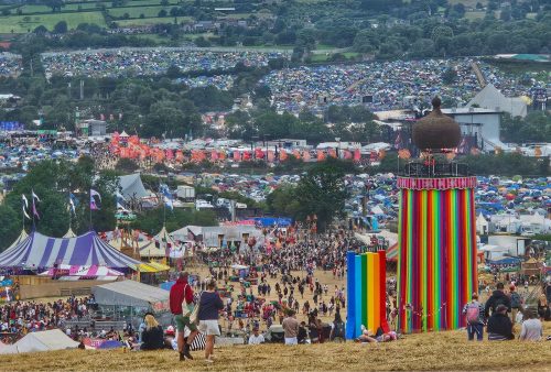 A large outdoor music festival with colorful tents, a striped tower, and crowds of people offers the perfect getaway. Many tents stretch across the green landscape under a cloudy sky, creating a vibrant scene for travel lovers.