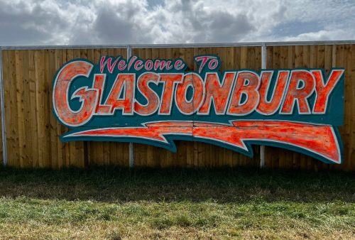 A wooden fence displays a large, colorful sign reading “Welcome to GLASTONBURY” in bold, red and turquoise letters, with lush grass and bush in the foreground—hinting at a vibrant travel getaway under a cloudy sky.