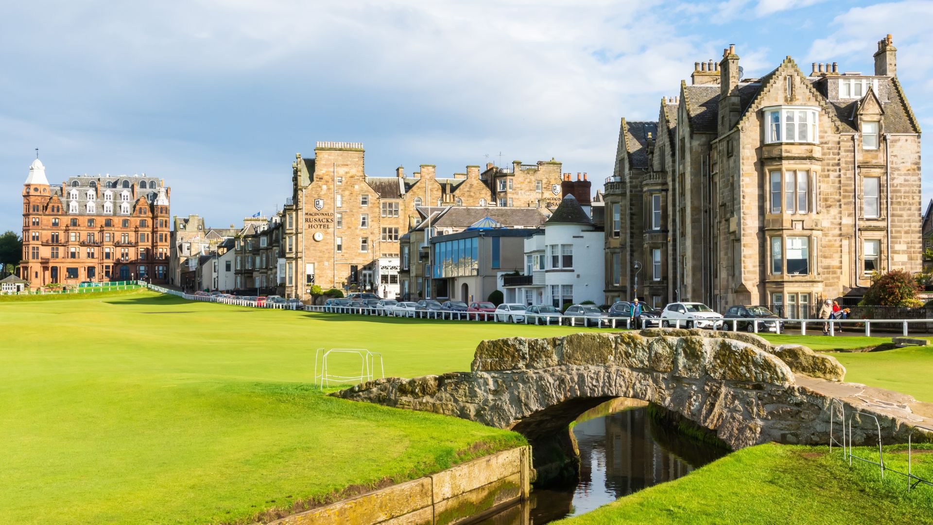 A stone bridge crosses a small stream on a golf course, with historic stone buildings and parked cars lining the street in the background under a partly cloudy sky—a perfect travel getaway surrounded by nature and the occasional bush.