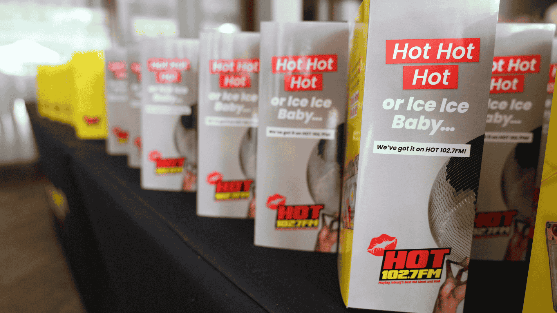 Several table tents featuring the HOT 102.7 FM logo and playful phrases like "Hot Hot Hot" or "Ice Ice Baby…" are lined up across a black-clothed table, setting the stage for a sizzling hot 1027 breakfast event.