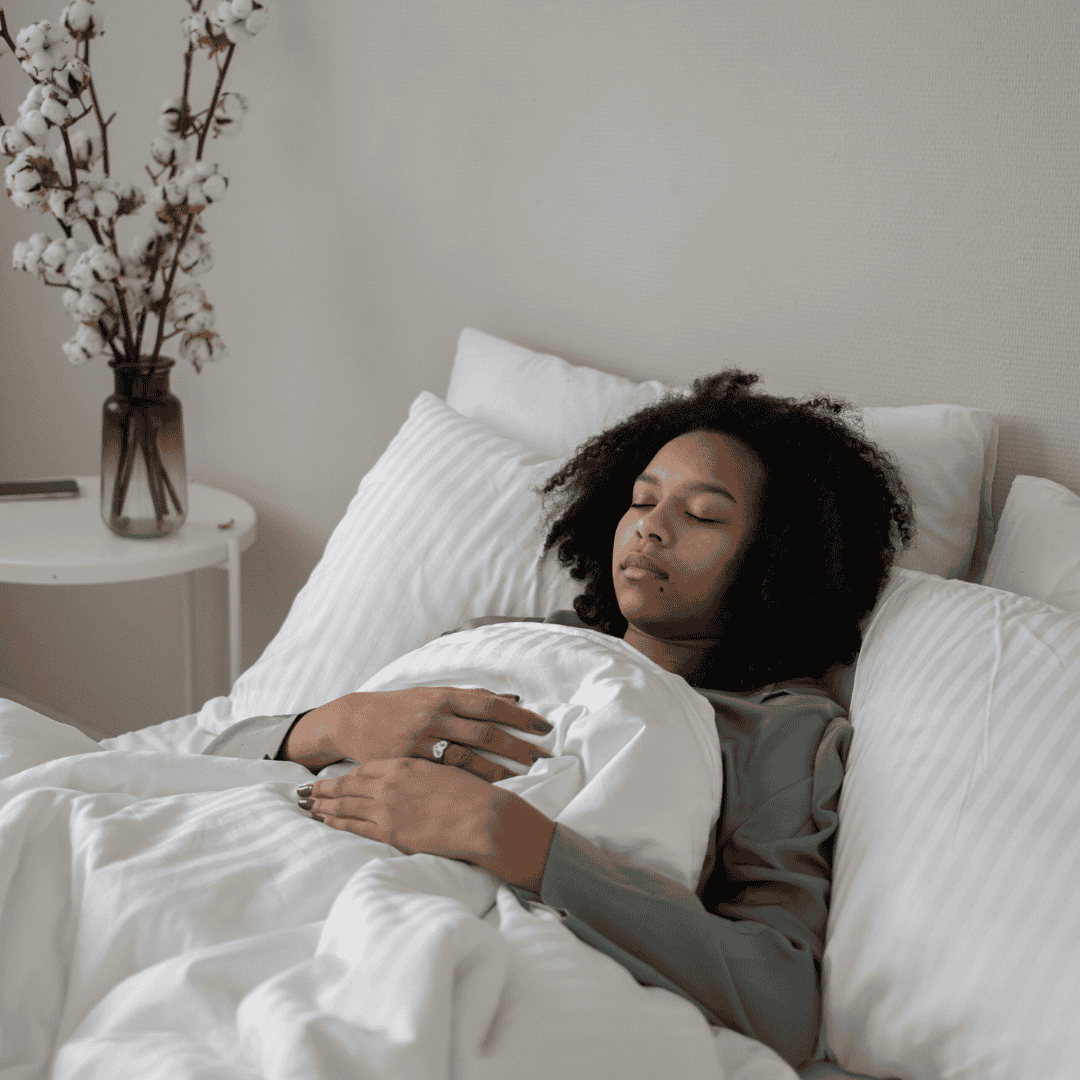 A woman with curly hair lies on her back in bed, resting with her eyes closed as sunlight streams in. White pillows and bedding surround her, and a vase of cotton stems sits on the bedside table—perfect relaxation before tuning into Hot 1027 Breakfast.