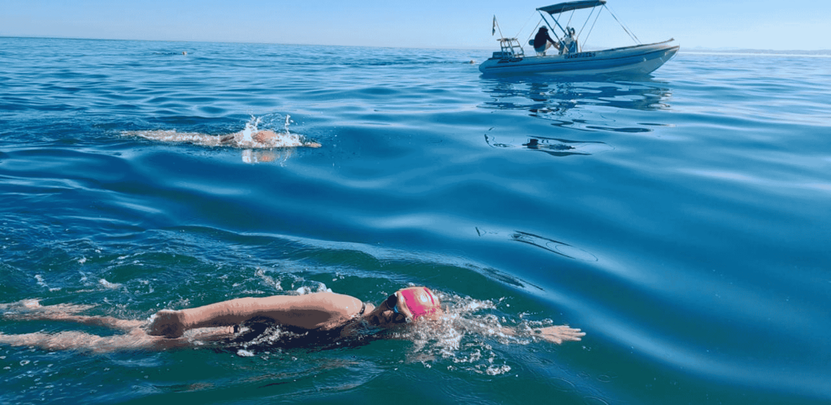 Two swimmers in open water, both wearing swim caps and goggles, swim near a small boat on a calm, sunny day. The boat with people onboard—perhaps filming for Pet TV—follows alongside on the gently rippling blue sea.