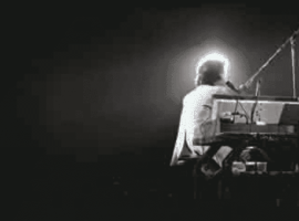 A person sits at a keyboard on a dark stage, illuminated from behind by a bright light, creating a glowing halo effect that hints at the wealth of talent and emotion in the dramatic silhouette of the musician.