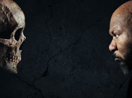 A man with a serious expression faces a human skull against a dark, cracked background, both in profile and staring intently at each other.