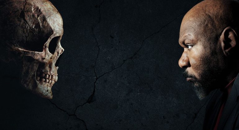 A man with a serious expression faces a human skull against a dark, cracked background, both in profile and staring intently at each other.
